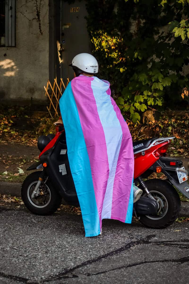 Person with a transgender pride flag draped over a scooter at night.
