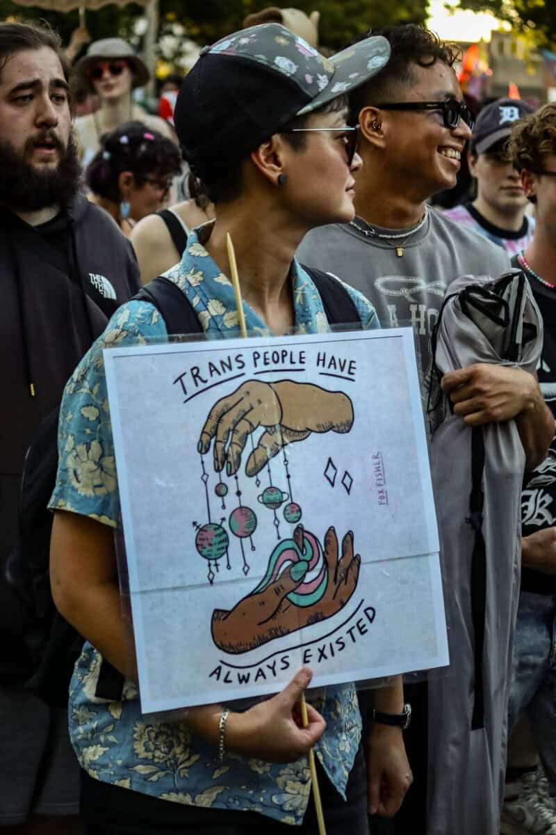 Trans march participant holding a sign with a hand-drawn illustration and message.