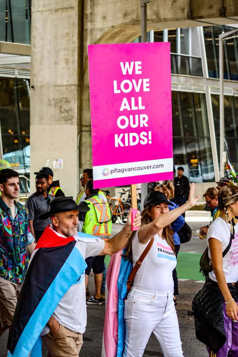 Protesters at a pride parade holding a pink sign that says "We Love All Our Kids!".