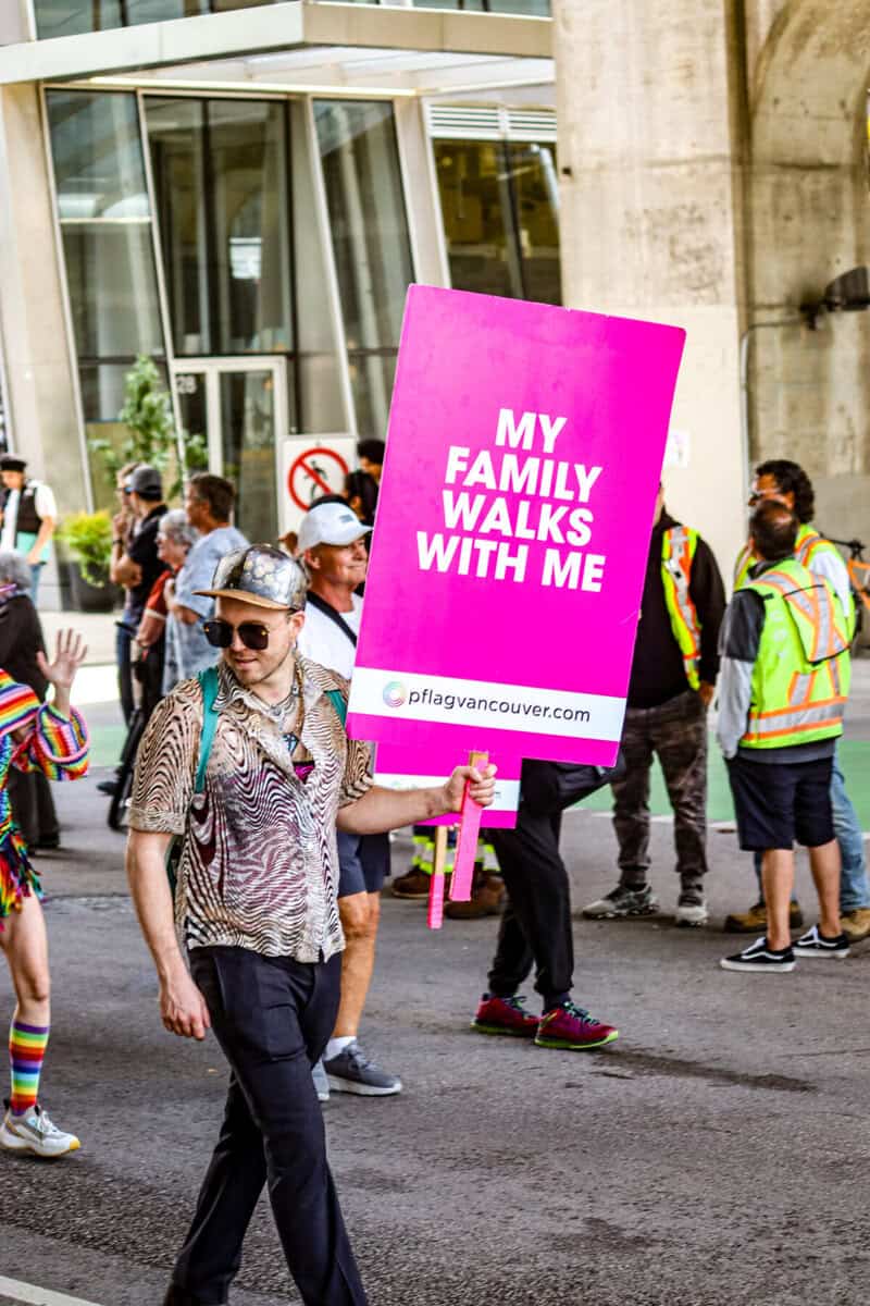 LGBTQ+ pride parade with diverse participants and a pink sign reading "My Family Walks With Me".
