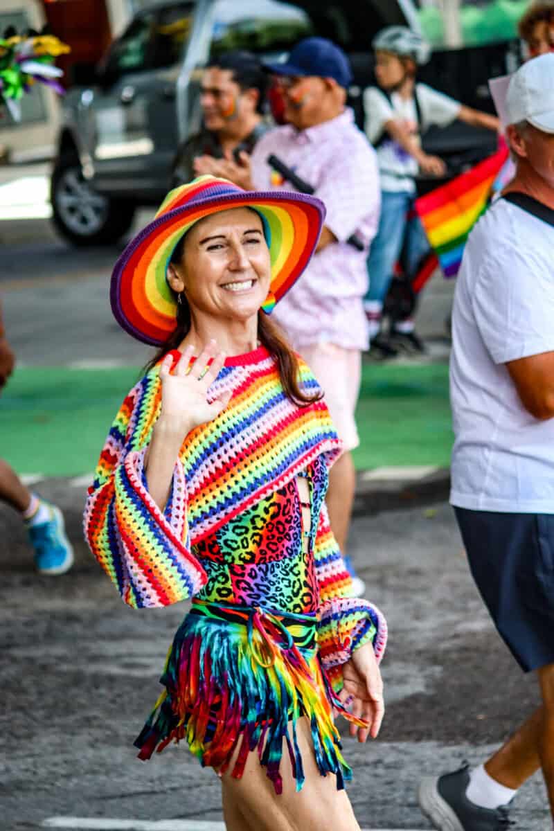 Woman smiling at Pride parade wearing rainbow hat and colorful outfit.
