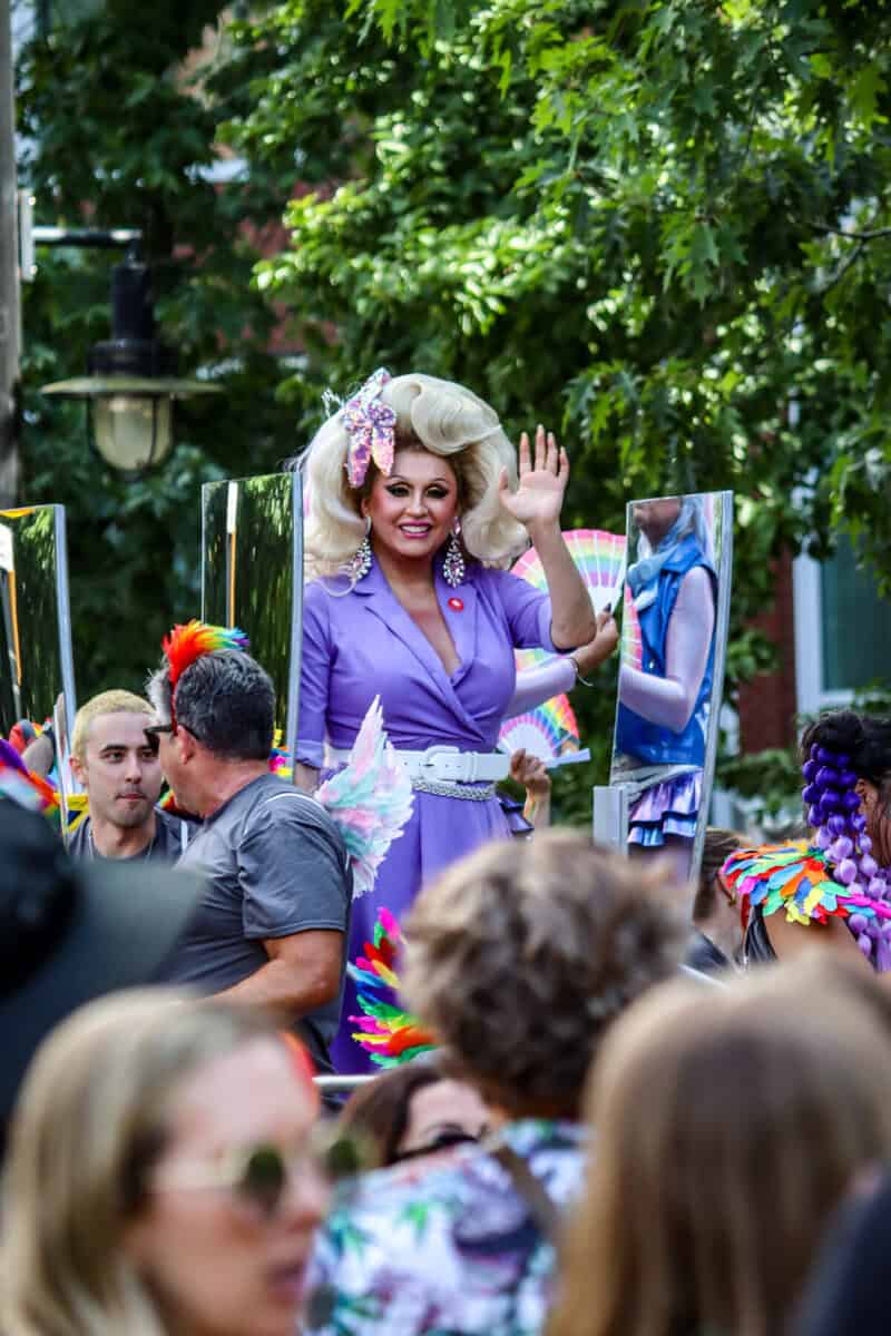 Drag queen in purple outfit waving at parade crowd with colorful costumes.