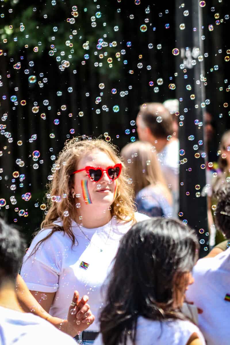 People celebrating Pride with rainbow accessories and bubbles outdoors in sunlight.