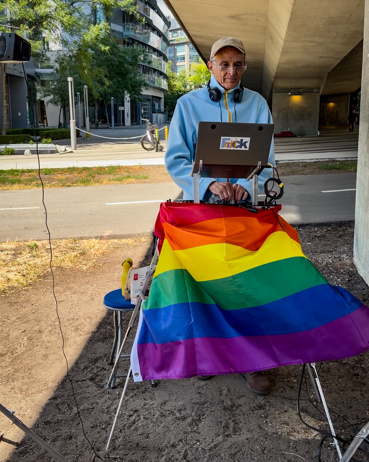 Older man with headphones and laptop at pride event with rainbow flag.