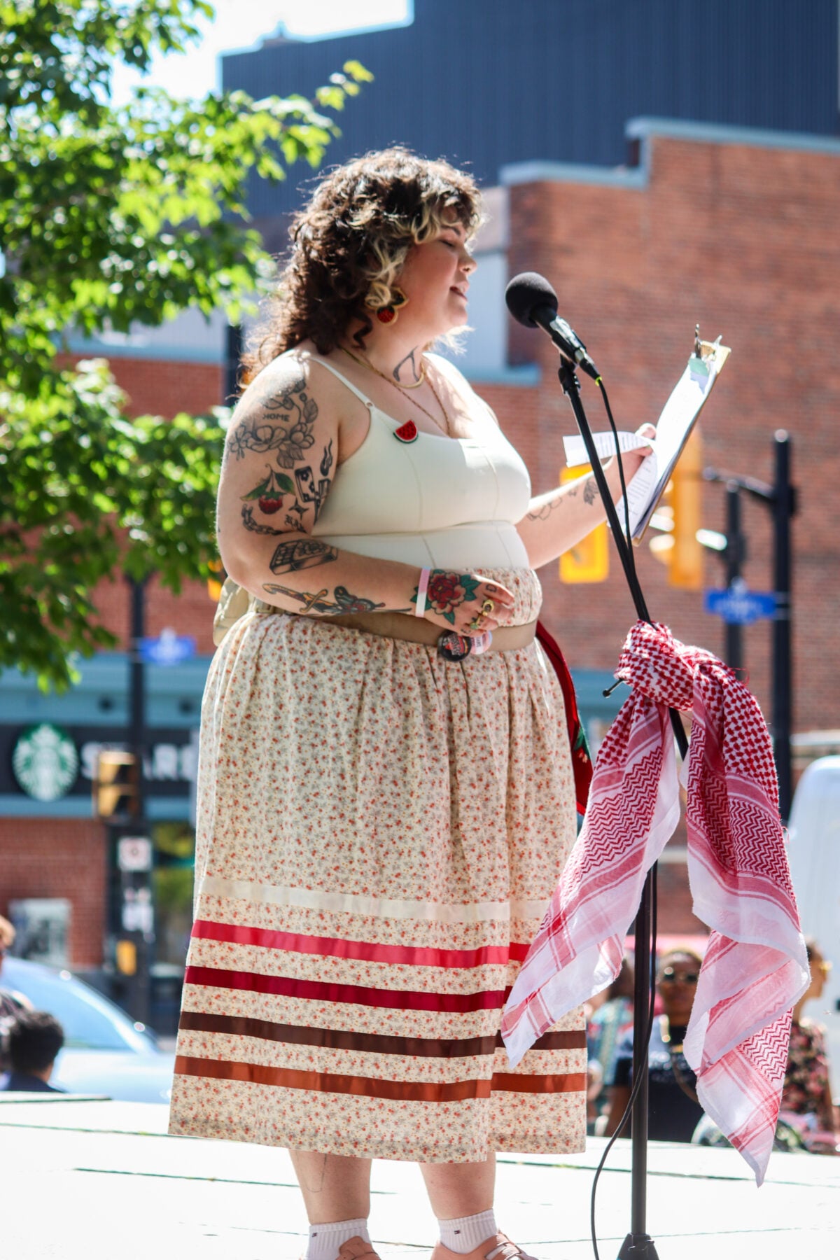 Woman speaking at outdoor event with microphone and red-and-white scarf.