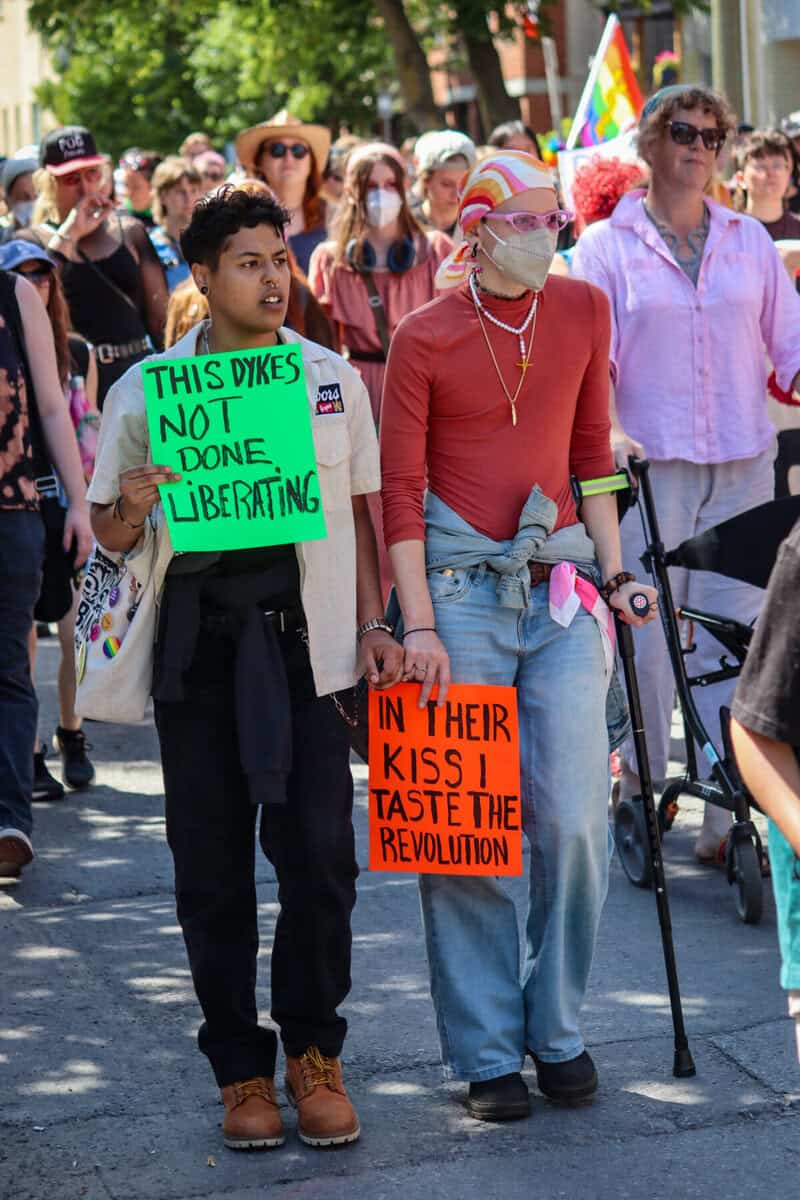Diverse group at pride march holding protest signs and rainbow flags.