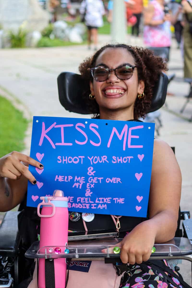 Woman in wheelchair holding a 'KISS ME' sign at outdoor event.