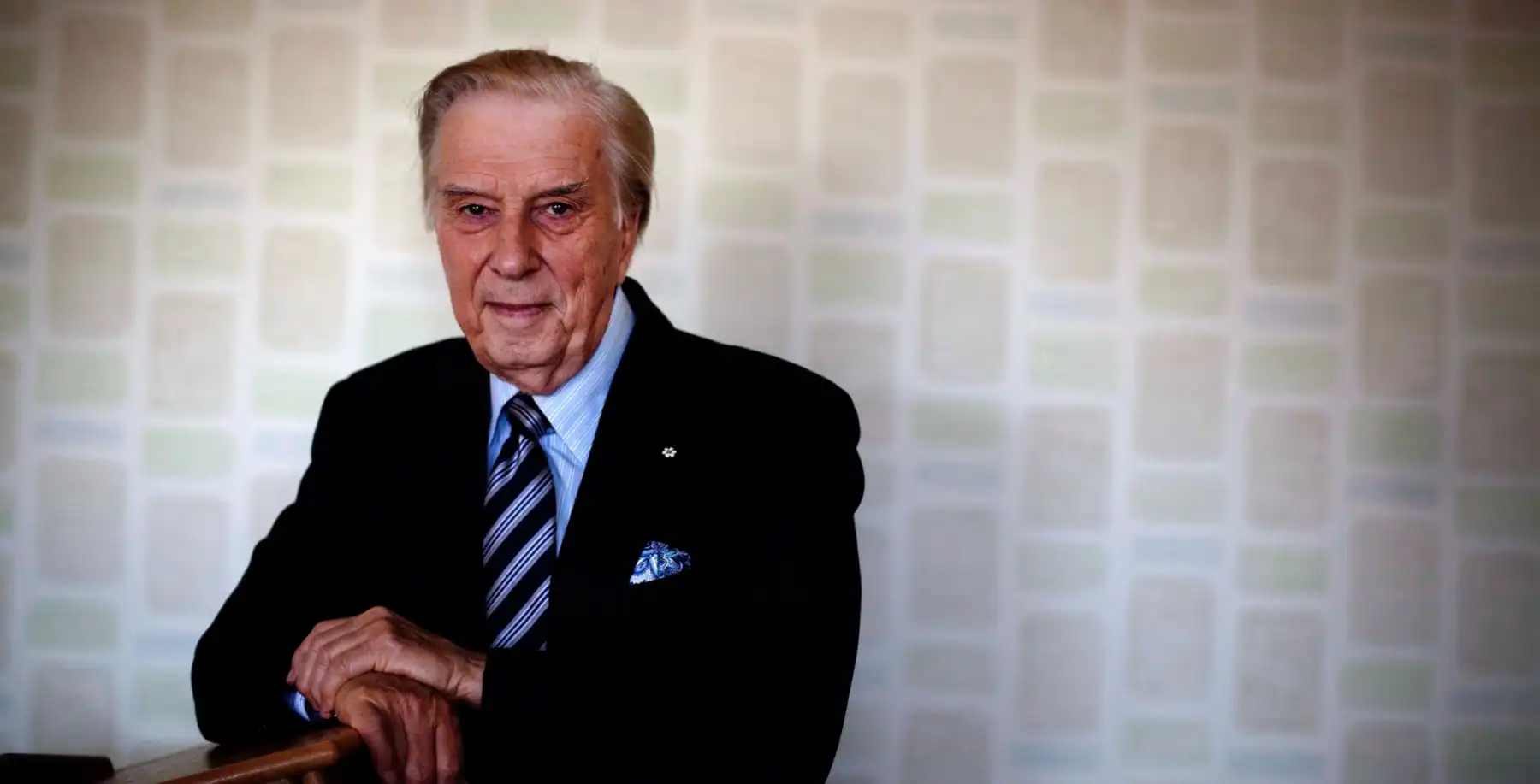 Elderly man in formal suit with a blue tie, standing indoors.