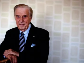 Elderly man in formal suit with a blue tie, standing indoors.